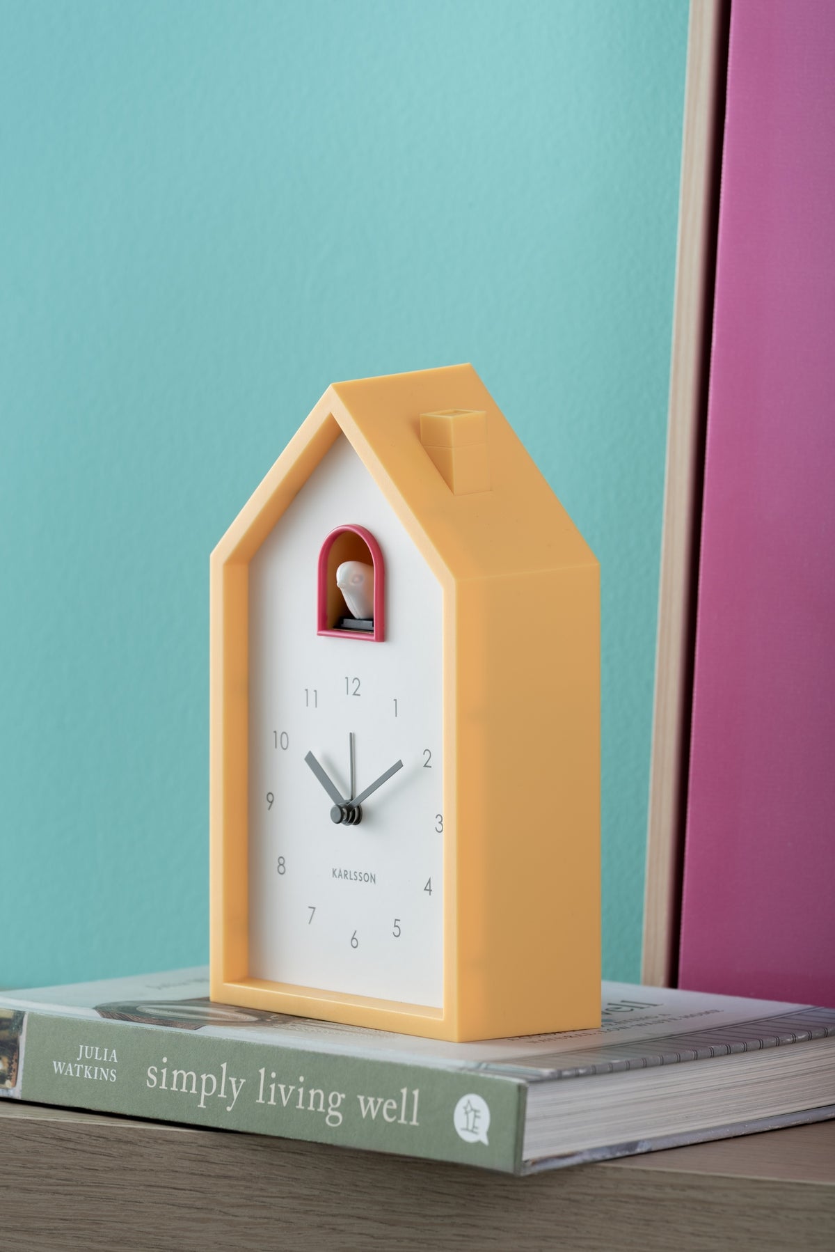 Karlsson wekker Modern Cuckoo, heldergeel, sfeerfoto: op boek tegen turquoise muur met roze paneel