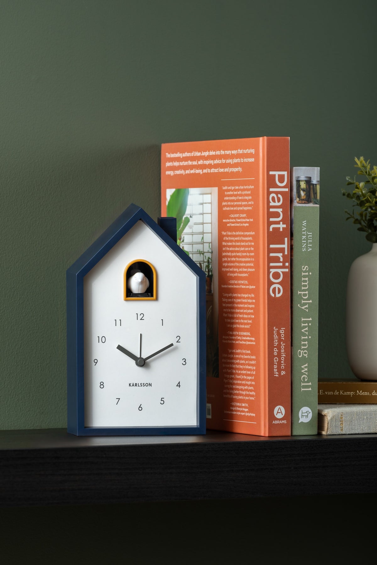 Karlsson wekker Modern Cuckoo, donkerblauw abs, sfeerfoto: op zwarte plank tegen groene muur naast boeken