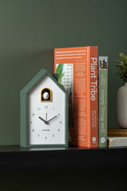 Karlsson wekker Modern Cuckoo, jungle groen abs, sfeerfoto: op zwarte plank tegen groene muur naast boeken
