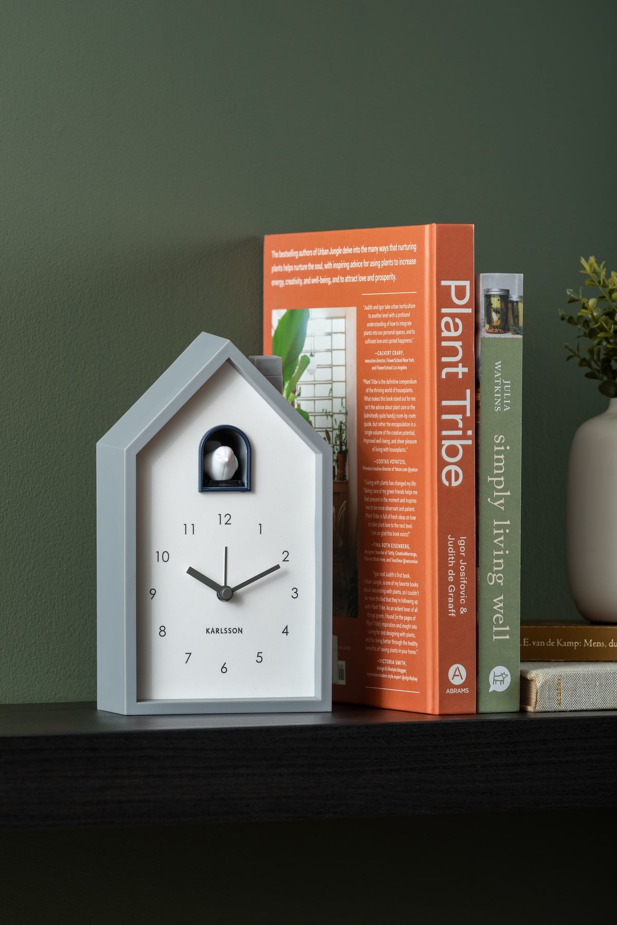 Karlsson wekker Modern Cuckoo, muisgrijs abs, sfeerfoto: op zwarte plank tegen groene muur naast boeken