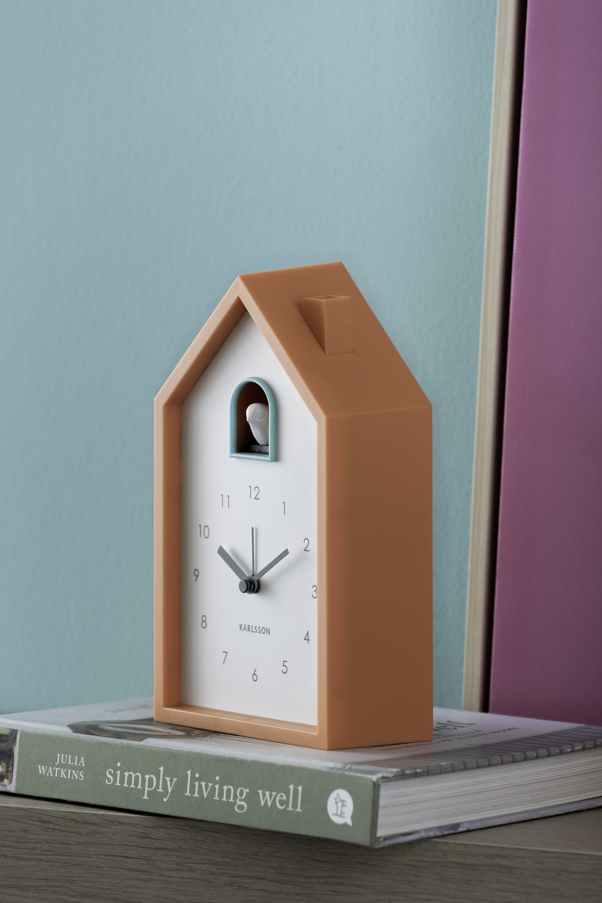 Karlsson wekker Modern Cuckoo, helderoranje, sfeerfoto: op boek tegen turquoise muur met roze paneel