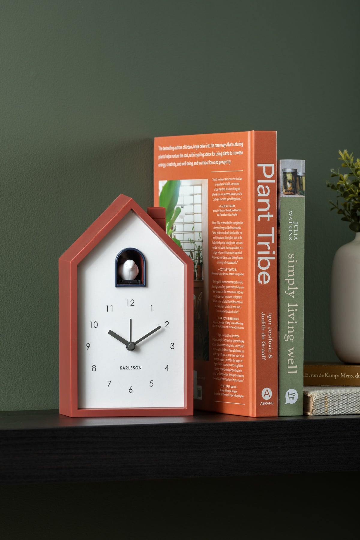 Karlsson wekker Modern Cuckoo, okerrood, sfeerfoto: op zwarte plank tegen groene muur naast boeken