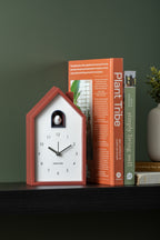 Karlsson wekker Modern Cuckoo, okerrood, sfeerfoto: op zwarte plank tegen groene muur naast boeken