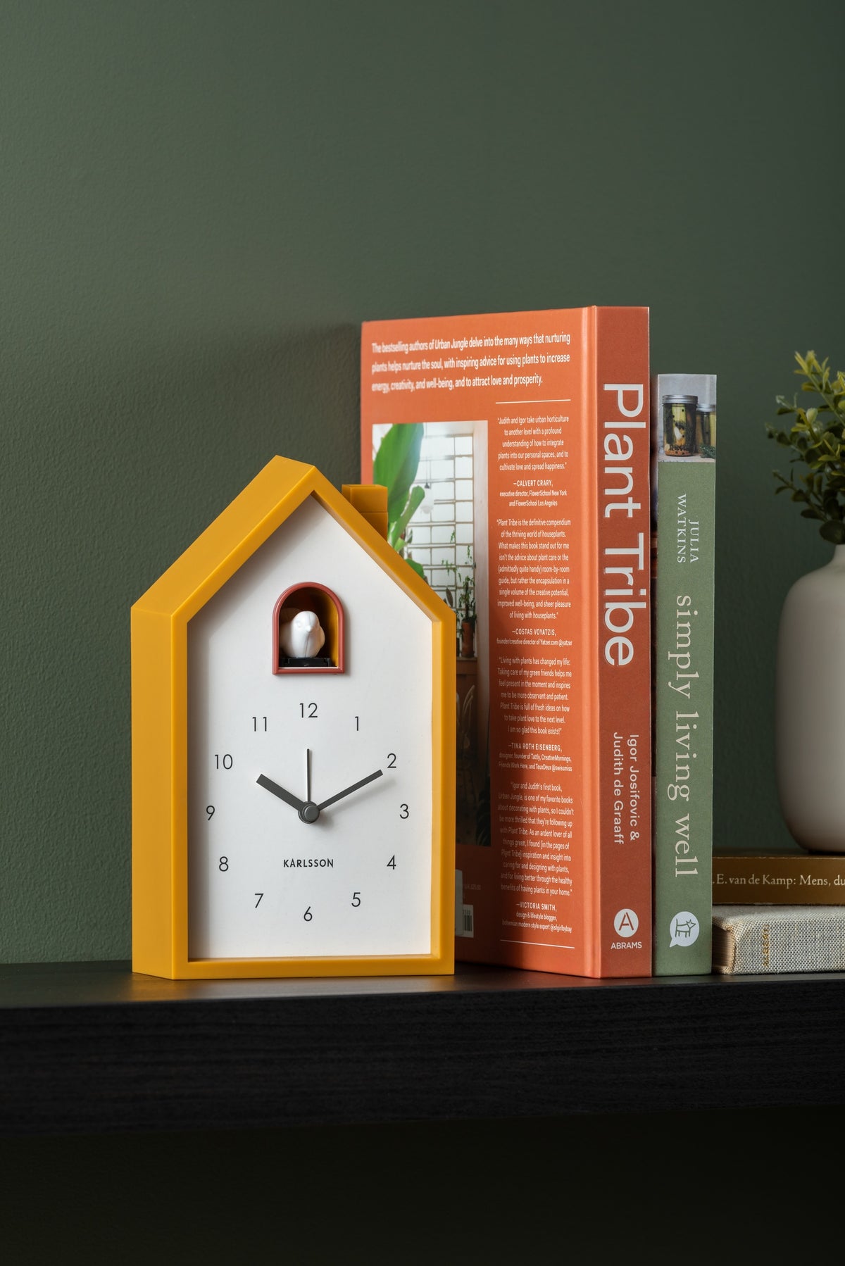 Karlsson wekker Modern Cuckoo, okergeel abs, sfeerfoto: op zwarte plank tegen groene muur naast boeken