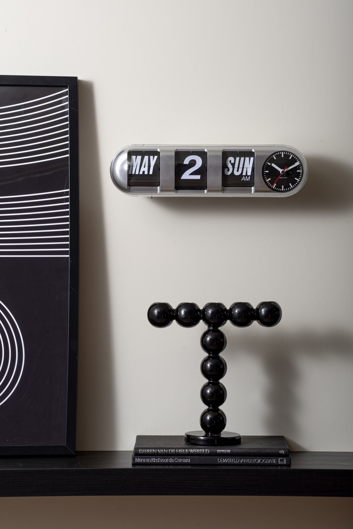Karlsson flipklok Tube Flip, zilver, sfeerfoto: aan wand boven zwarte kaarsenstandaard en boeken, naast kunstwerk