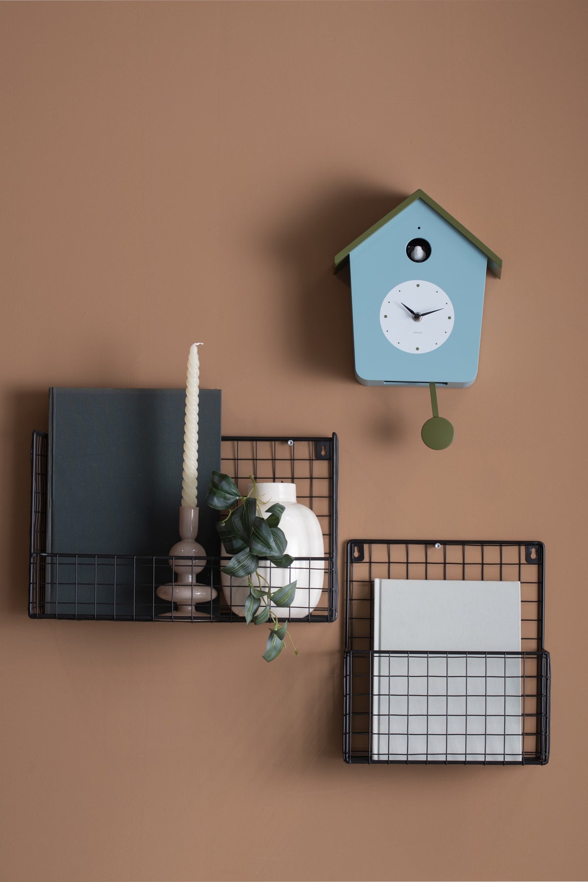Karlsson koekoeksklok Cuckoo Light, mistig blauw en legergroen, sfeerfoto: aan terracotta wand boven zwarte draadmanden