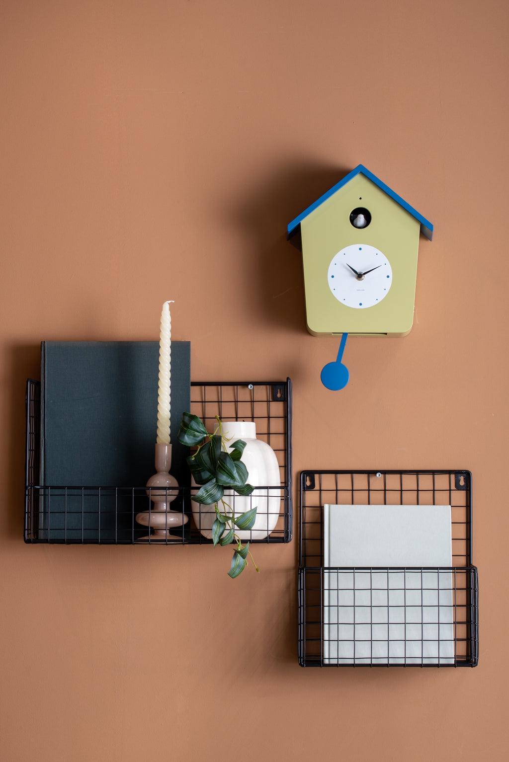Karlsson koekoeksklok Cuckoo Light, theegroen en klassiek blauw, sfeerfoto: aan terracotta wand boven zwarte draadmanden