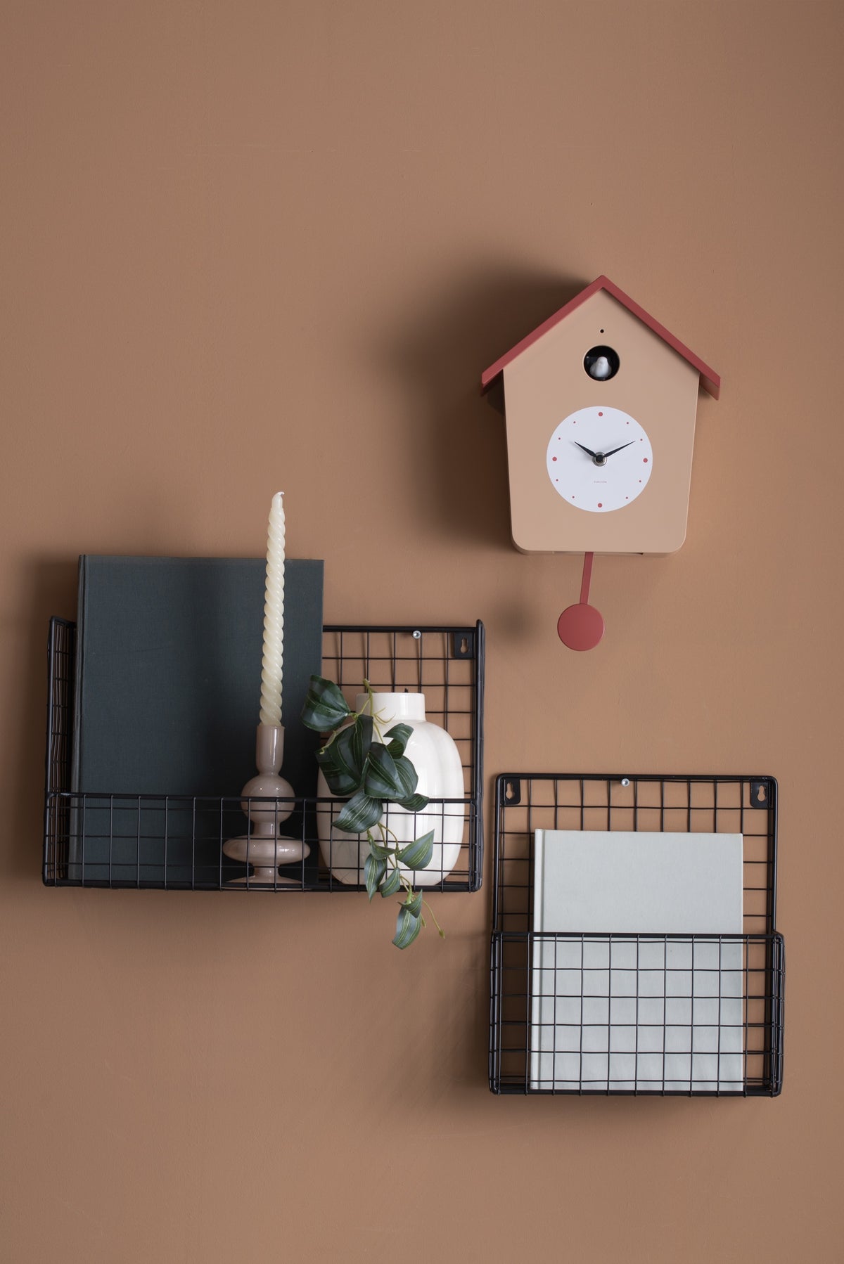 Karlsson koekoeksklok Cuckoo Light, zacht bruin en okerrood, sfeerfoto: aan terracotta wand boven zwarte draadmanden