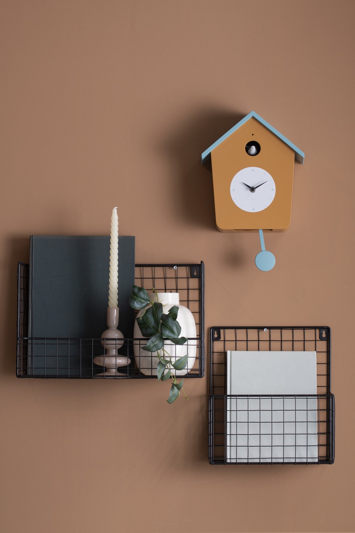 Karlsson koekoeksklok Cuckoo Light, okergeel en mistig blauw, sfeerfoto: aan terracotta wand boven zwarte draadmanden