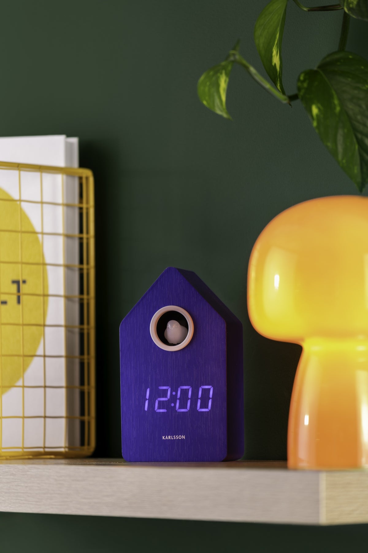 Karlsson wekker Cuckoo LED, blauw, sfeerfoto: op houten plank tussen gele mand en oranje lamp, groene muur, display 12:00