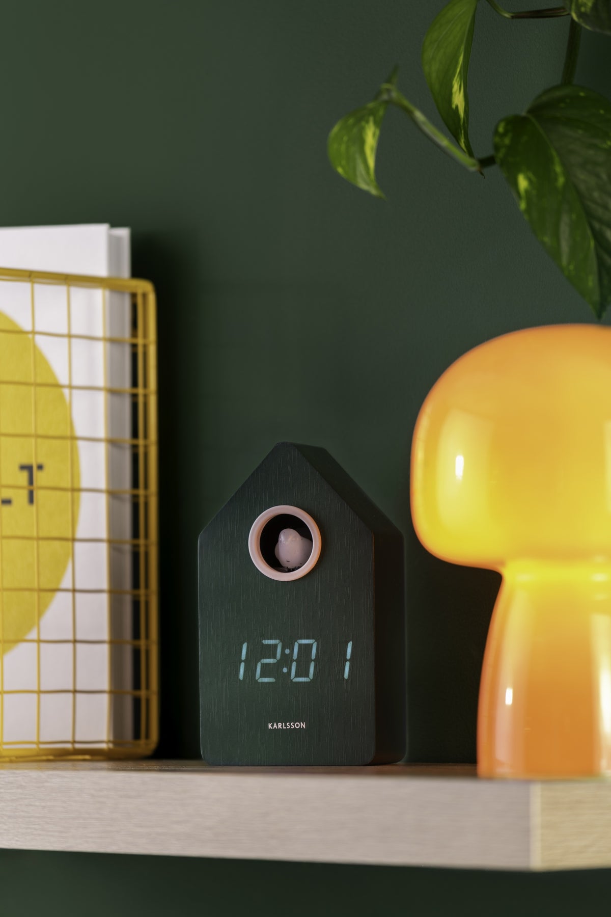 Karlsson wekker Cuckoo LED, groen, sfeerfoto: op houten plank tussen gele mand en oranje lamp, groene muur, display 12:01