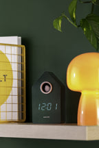 Karlsson wekker Cuckoo LED, groen, sfeerfoto: op houten plank tussen gele mand en oranje lamp, groene muur, display 12:01