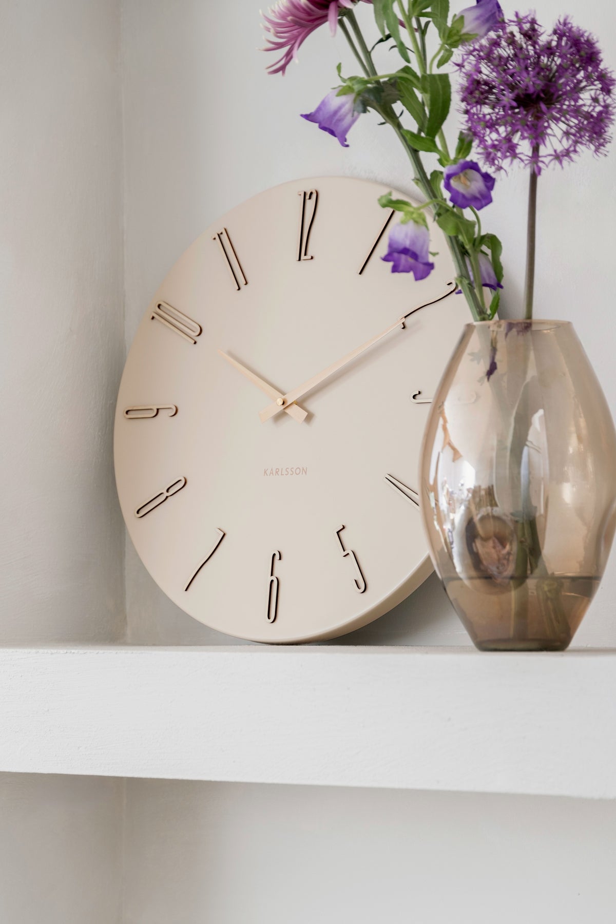 Karlsson wandklok Esmerado, wit, sfeerfoto: op witte plank tegen muur naast glazen vaas met paarse bloemen