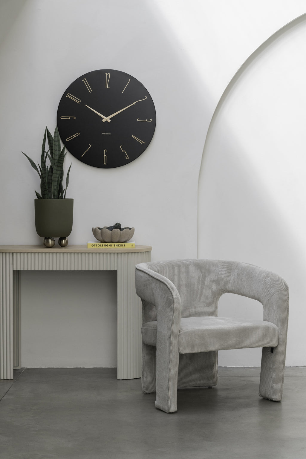 Karlsson wandklok Esmerado Large, zwart, sfeerfoto: aan witte muur boven sidetable met plant, naast grijze fauteuil