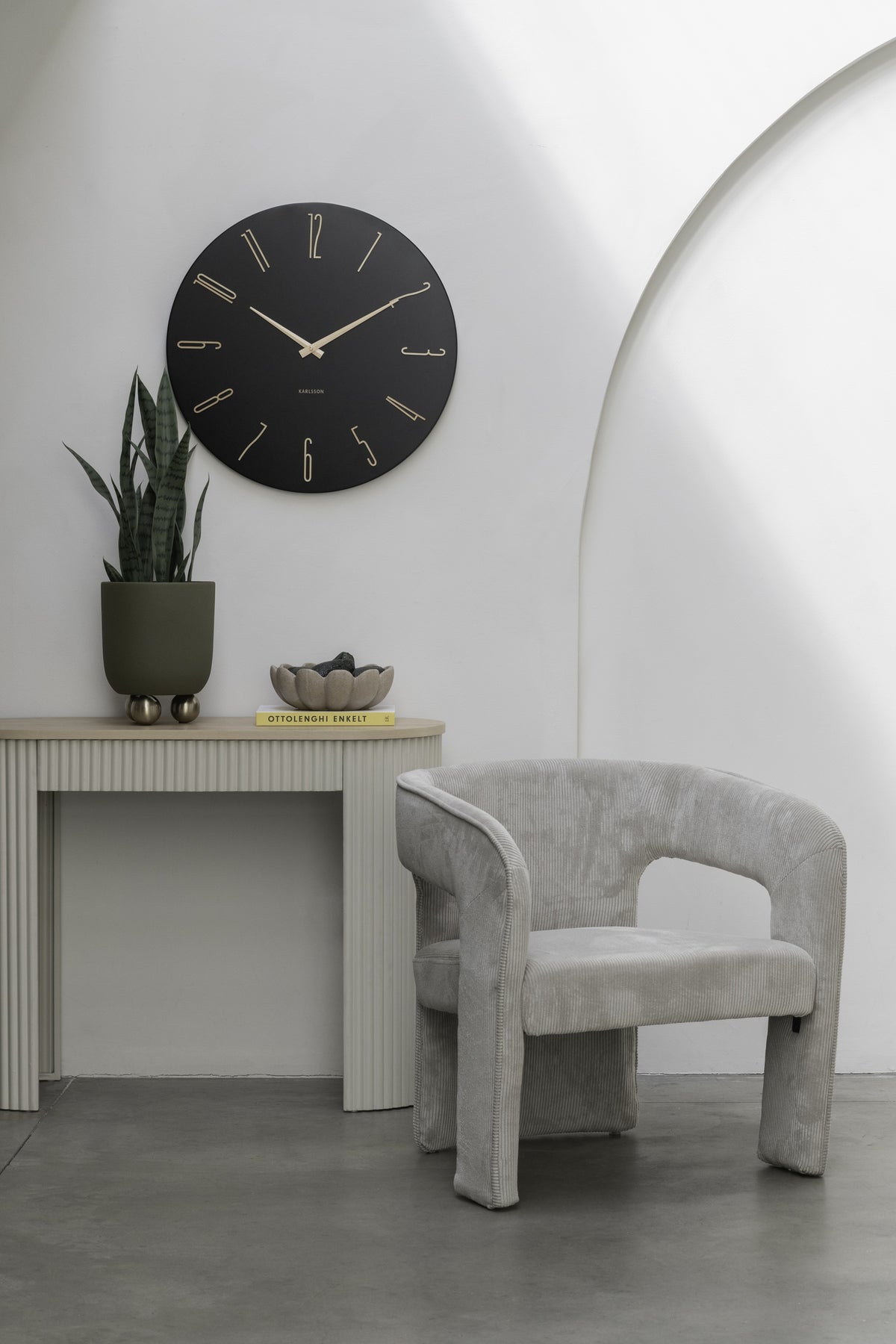 Karlsson wandklok Esmerado Large, zwart, sfeerfoto: aan witte muur boven sidetable met plant, naast grijze fauteuil