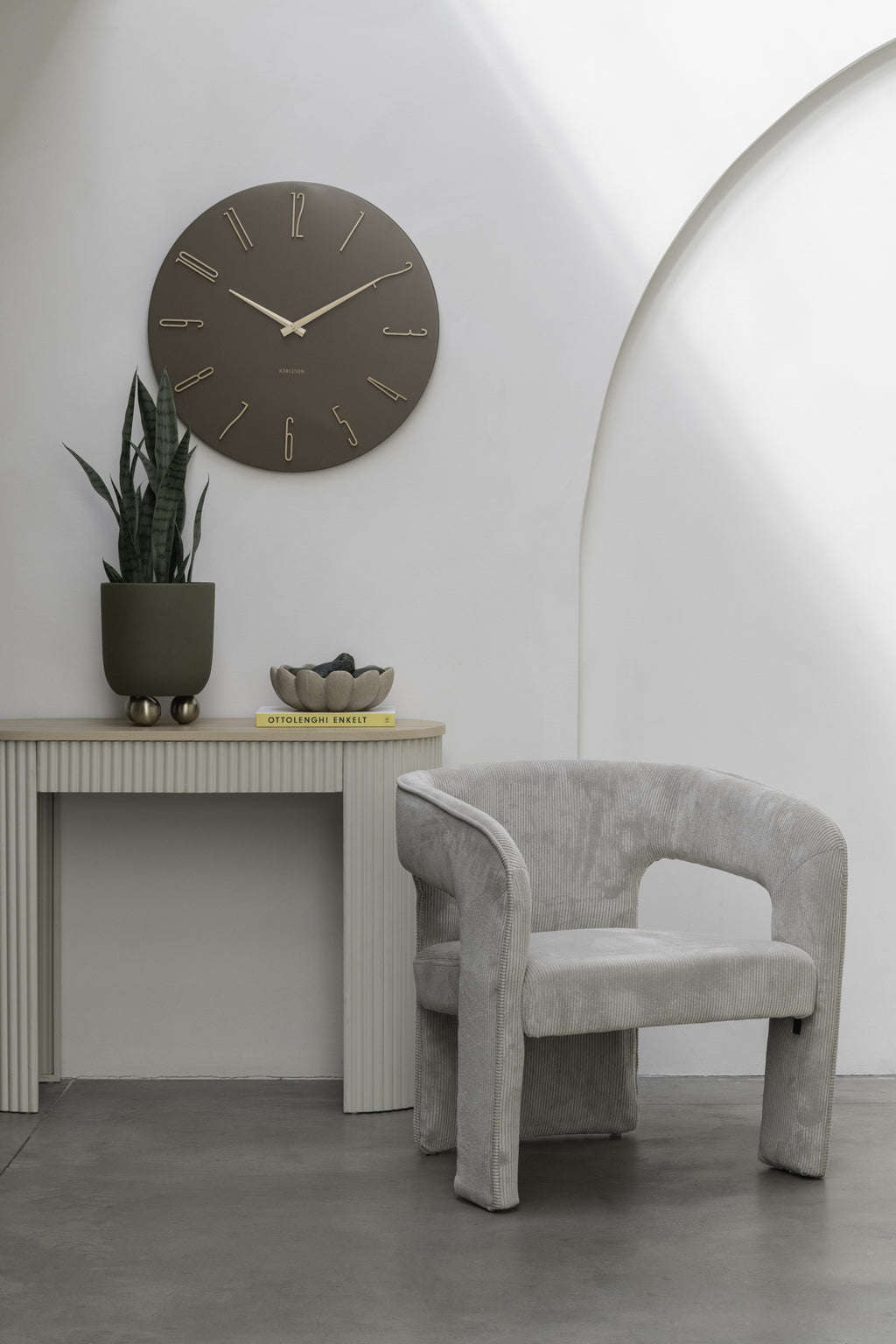 Karlsson wandklok Esmerado Large, bruin, sfeerfoto: aan witte muur boven sidetable met plant, naast grijze fauteuil