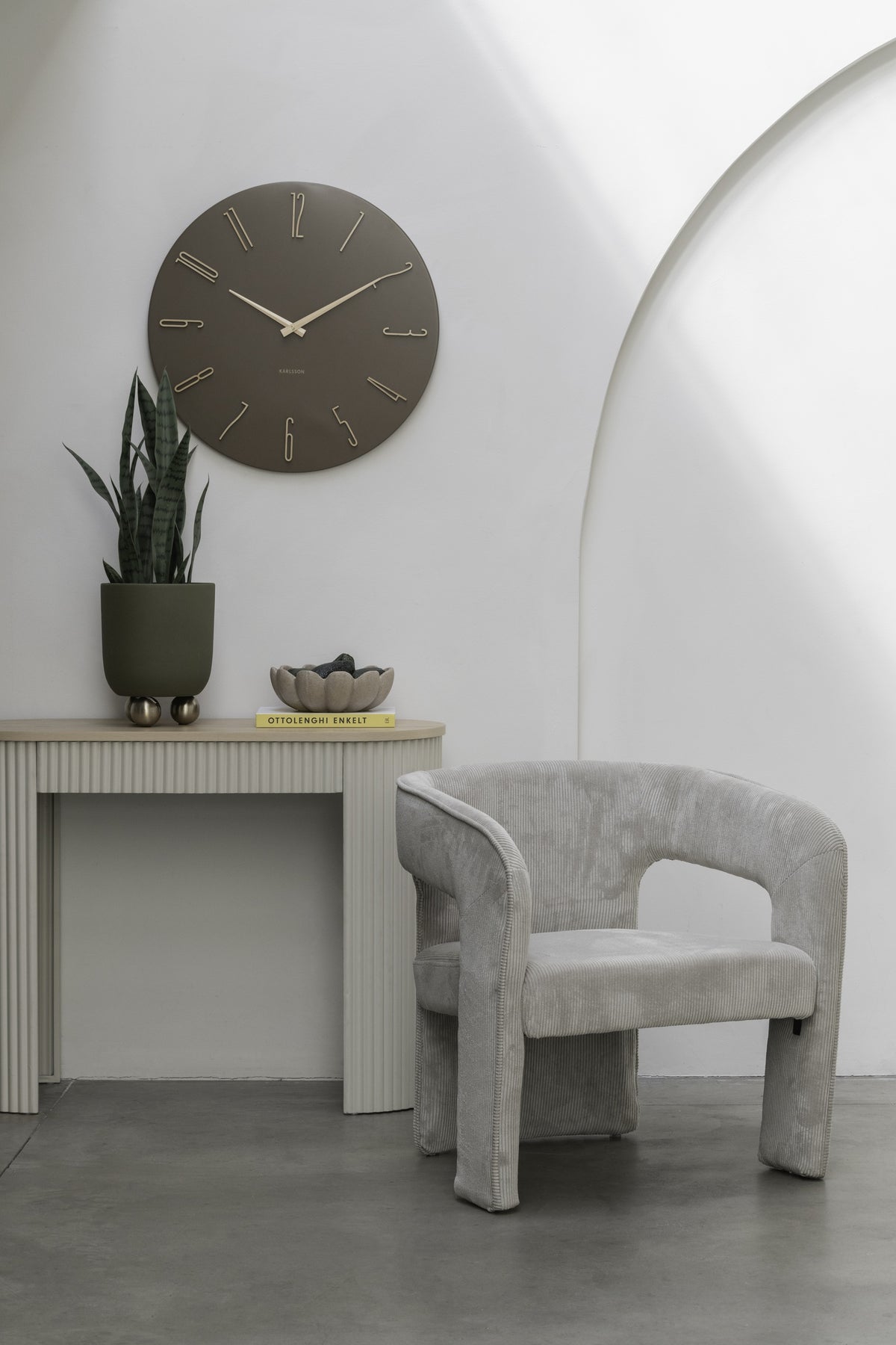 Karlsson wandklok Esmerado Large, bruin, sfeerfoto: aan witte muur boven sidetable met plant, naast grijze fauteuil