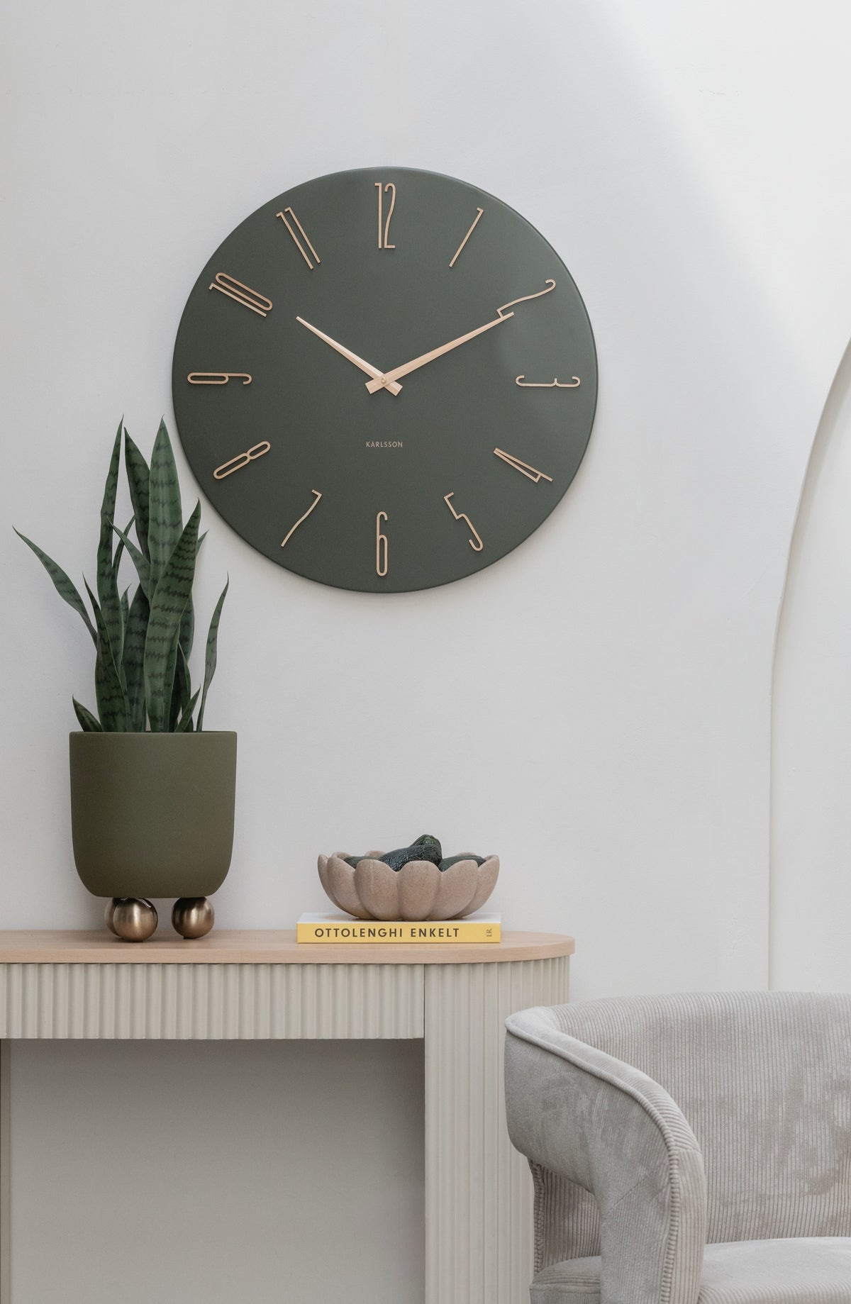 Karlsson wandklok Esmerado Large, groen, sfeerfoto: aan witte muur boven sidetable met plant en schaal, rand van fauteuil