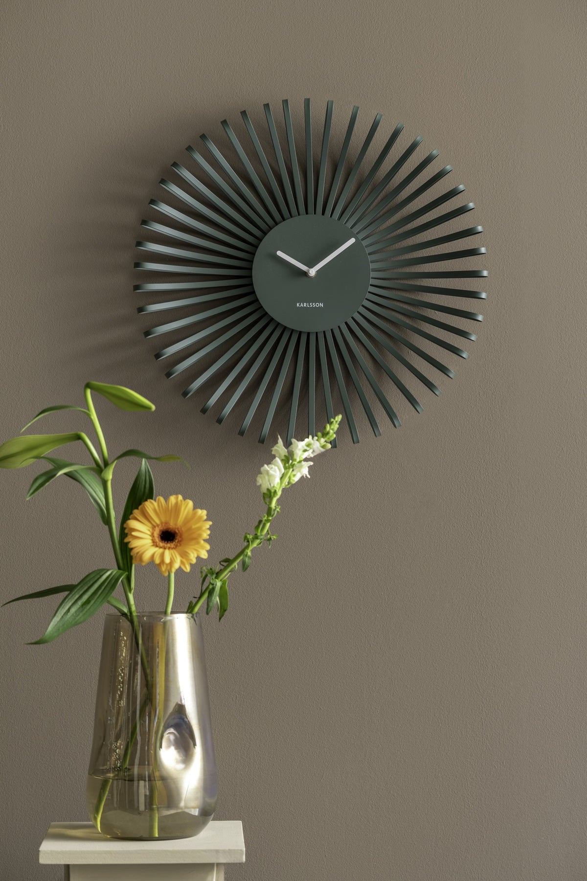 Karlsson wandklok Polo, groen, sfeerfoto: aan taupe muur boven glazen vaas met oranje gerbera