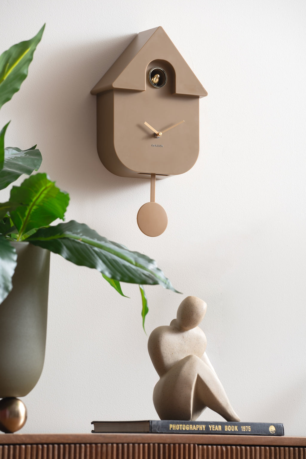 Karlsson koekoeksklok Modern Cuckoo Oro, mokka bruin, sfeerfoto: aan witte muur boven plant en beeldje op boek