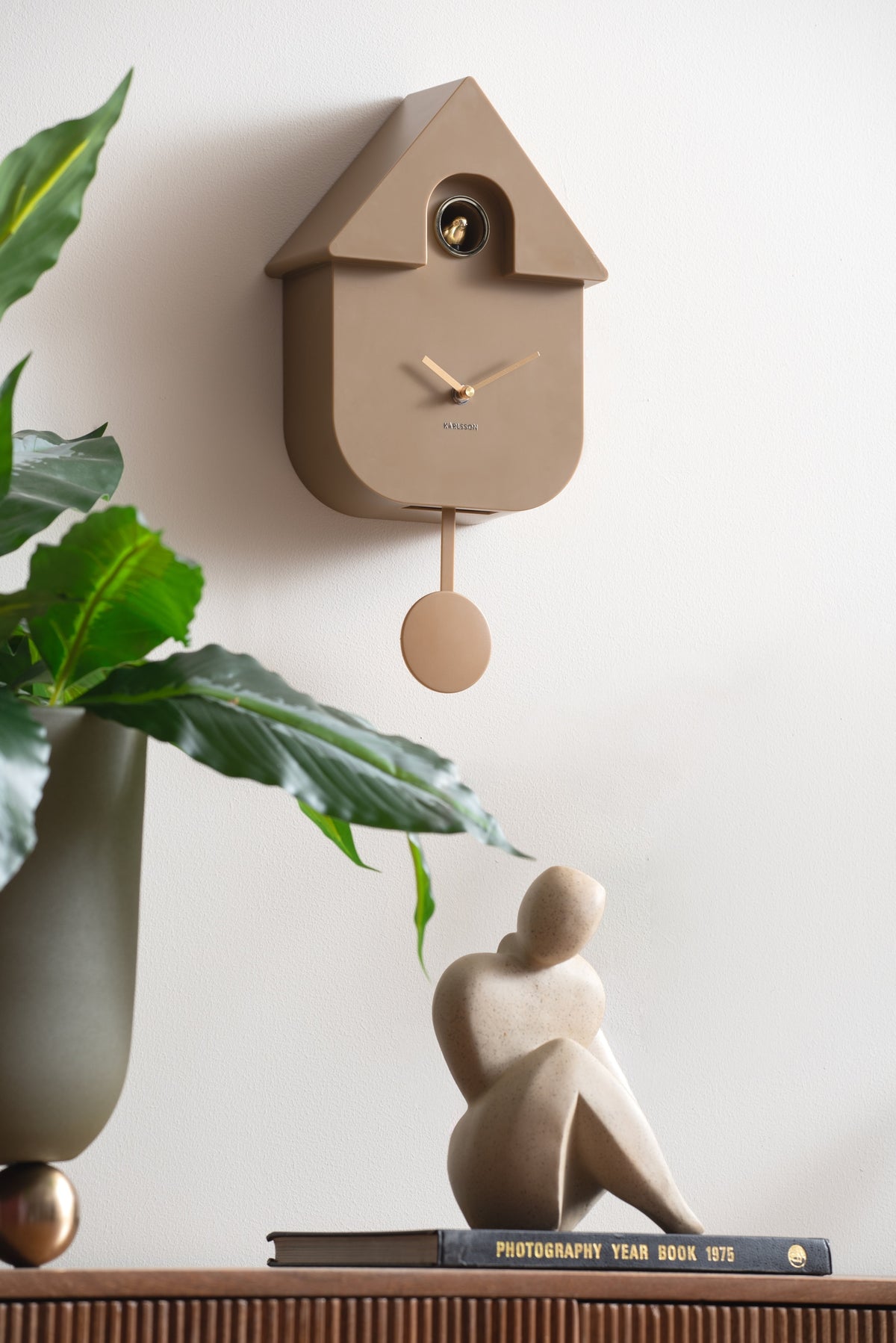 Karlsson koekoeksklok Modern Cuckoo Oro, mokka bruin, sfeerfoto: aan witte muur boven plant en beeldje op boek