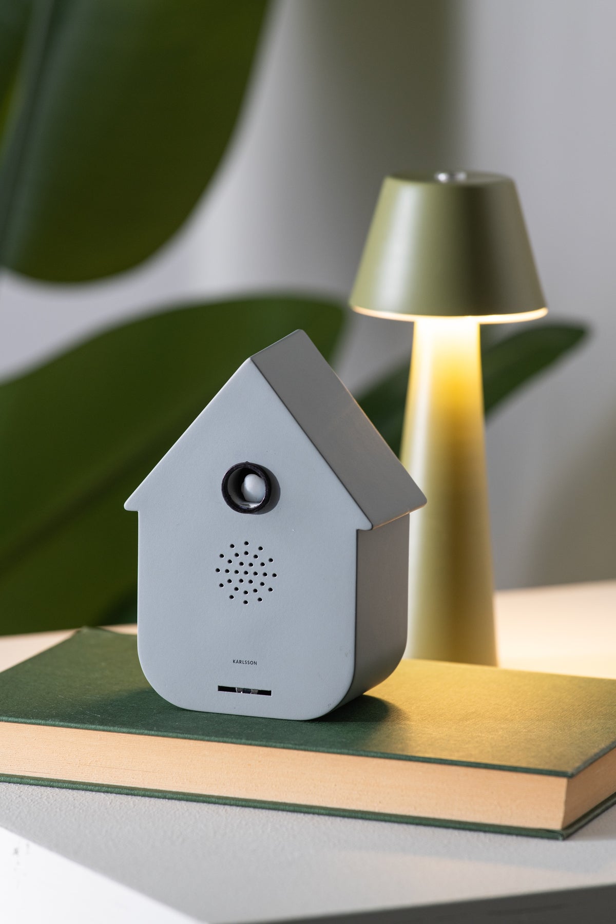 Karlsson vogelhuisje met geluid (soundbox) Modern Cuckoo, grijs, sfeerfoto: op groen boek naast tafellamp en plant