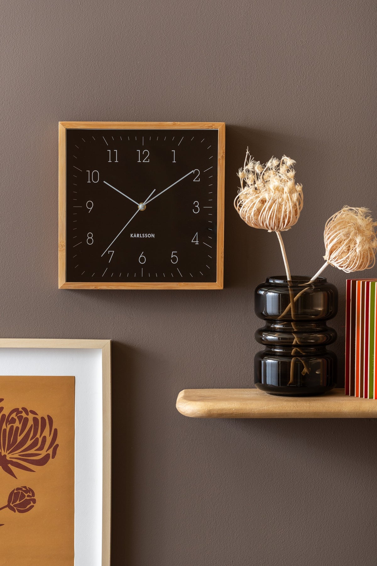 Karlsson wandklok Neat Cuadrado, zwart, sfeerfoto: aan bruine wand naast houten plankje met zwarte vaas en droogbloemen