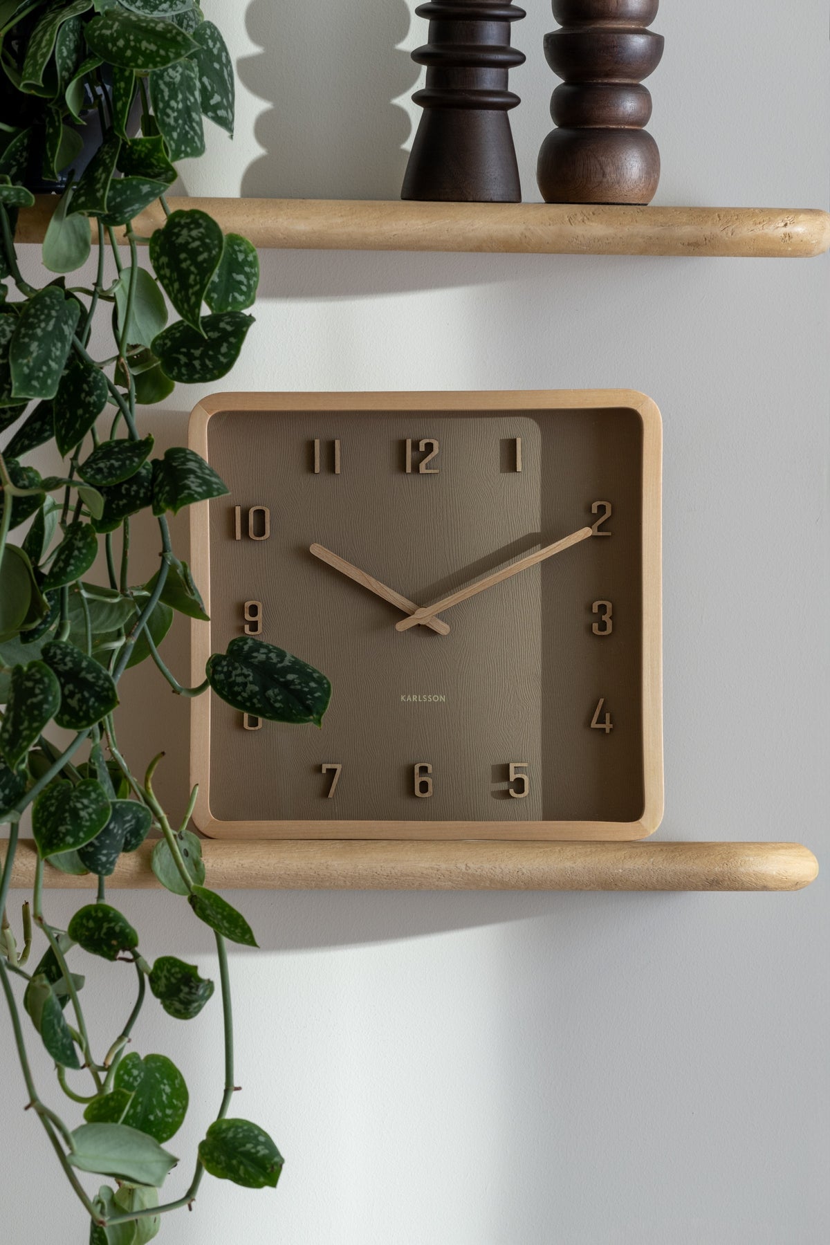 Karlsson wandklok Nervadura Square, zachtbruin en licht hout, sfeerfoto: op houten wandplank tussen hangplant en houten